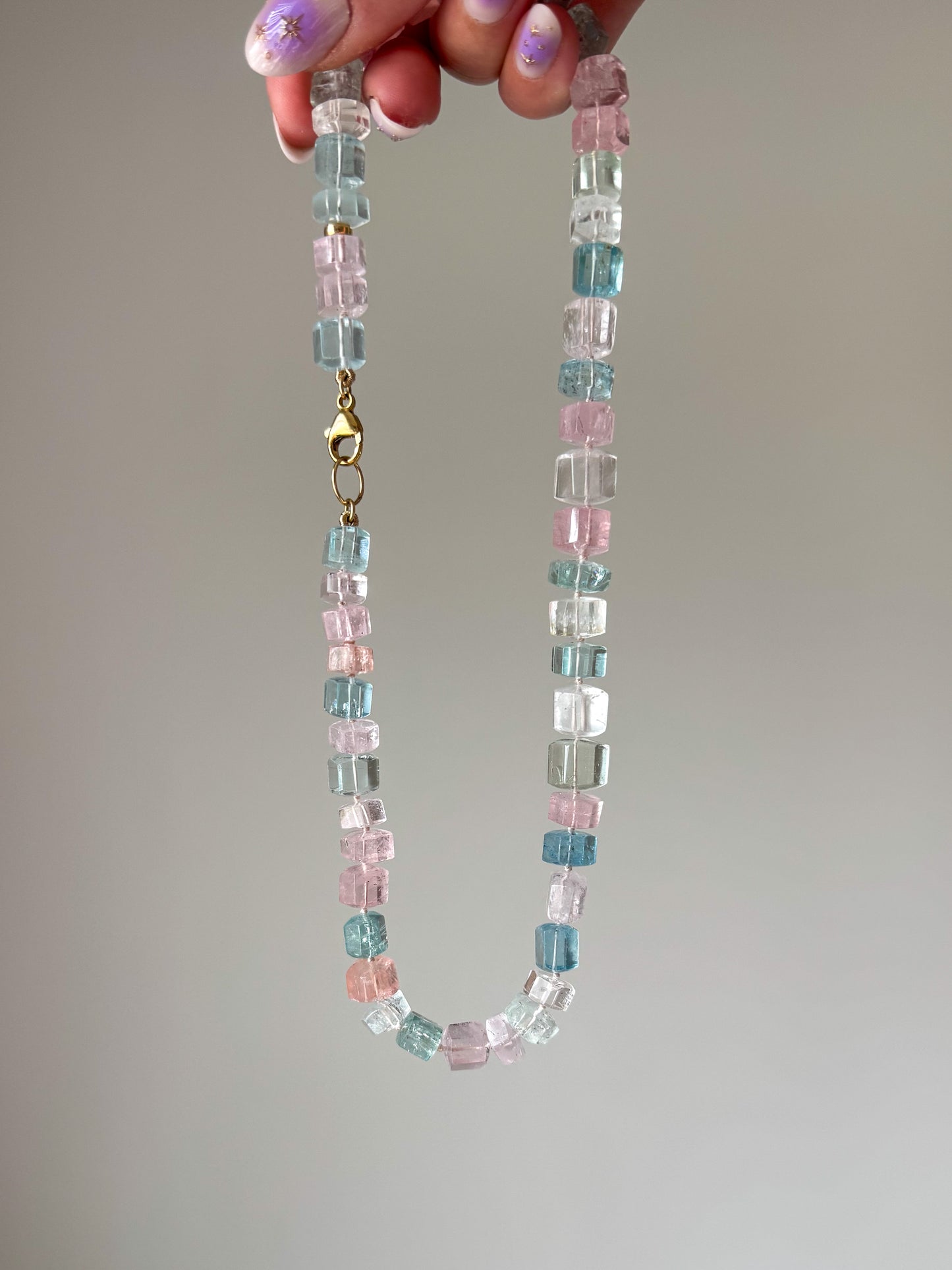Necklace with multicolored beads held by a hand on a neutral background aquamarine pink blue green peach white 14k gold
