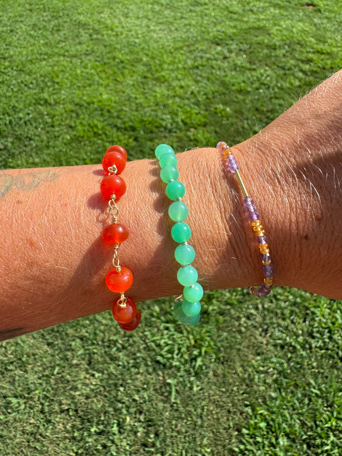 Three beaded bracelets on a wrist against a grass background carnelian chrysoprase 18k gold 14k yellow gold rose de France amethyst mandarin garnet