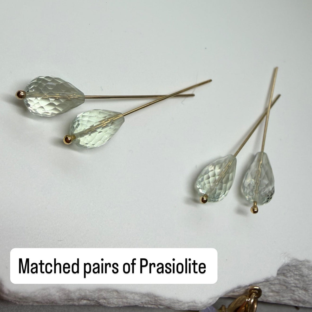 Four pairs of gold-tone stud earrings with faceted clear prasiolite gemstones, arranged on a white textured stone slab with a gray surface below. A text label reads 'Matched pairs of Prasiolite'.