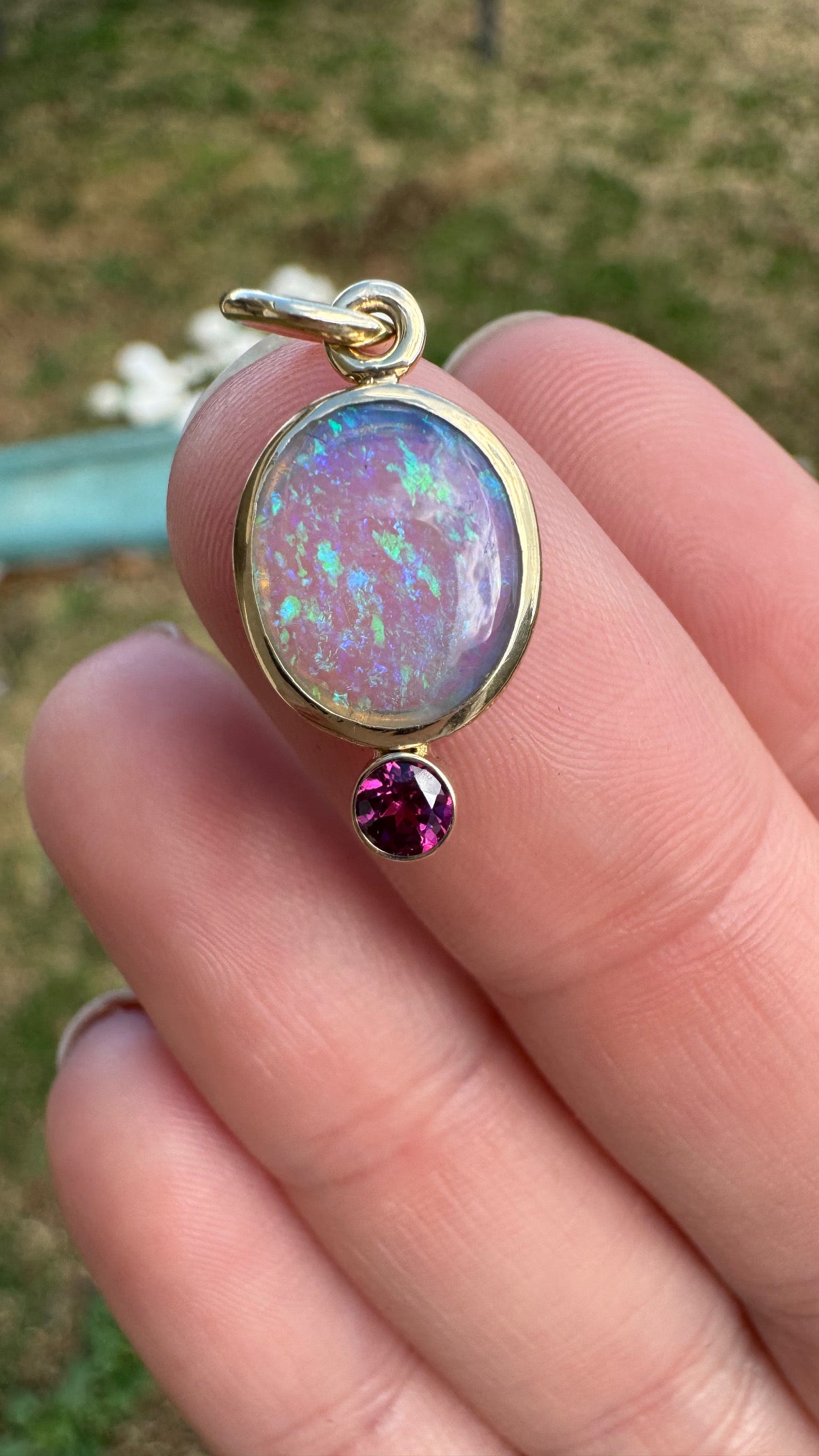 Opal pendant with a pink gemstone held between fingers against a blurred natural background