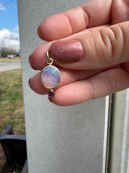 Opal and purple gemstone charm held between fingers with a blurred outdoor background