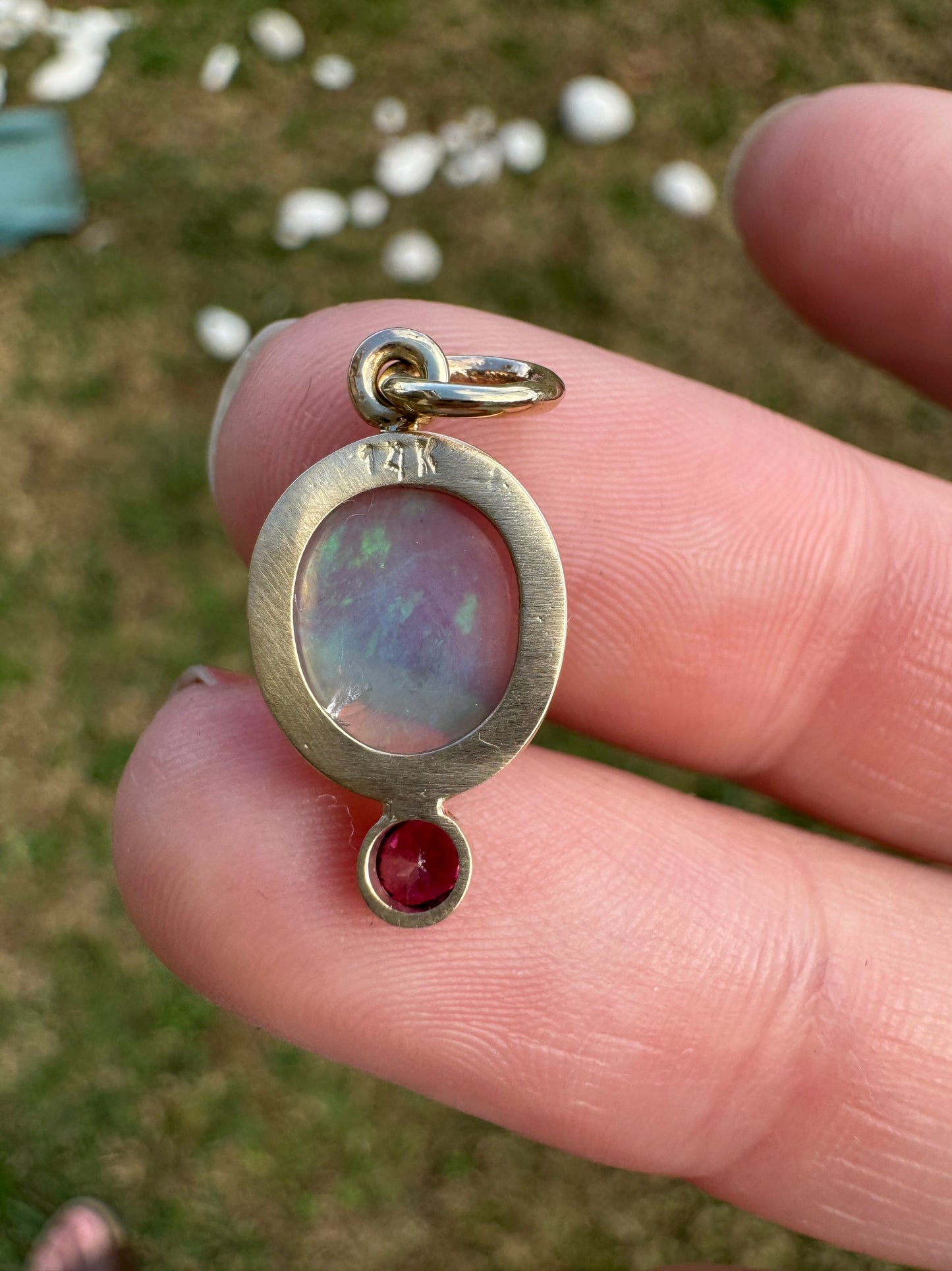 Gold pendant with an opal and red gemstone held between fingers against a blurred natural background