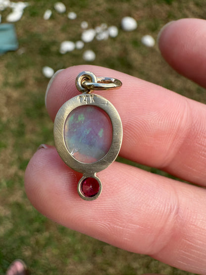 Gold pendant with an opal and red gemstone held between fingers against a blurred natural background