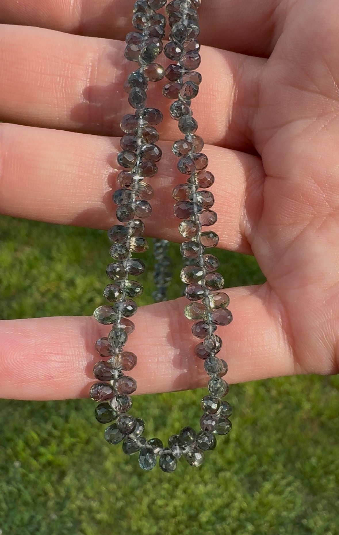 A close-up of a hand holding a teardrop-shaped green sapphire necklace, with each bead visible, set against a blurred background of grass.