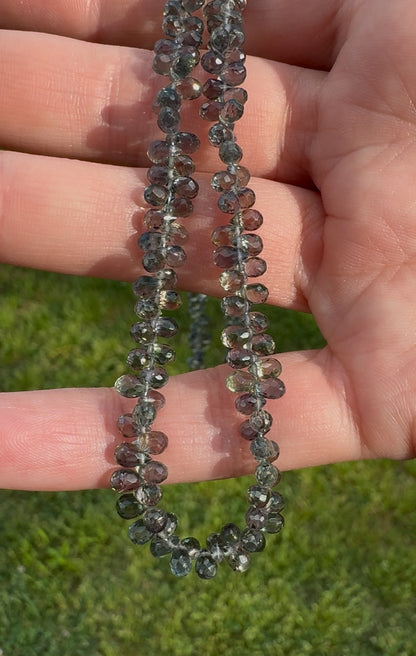 A close-up of a hand holding a teardrop-shaped green sapphire necklace, with each bead visible, set against a blurred background of grass.