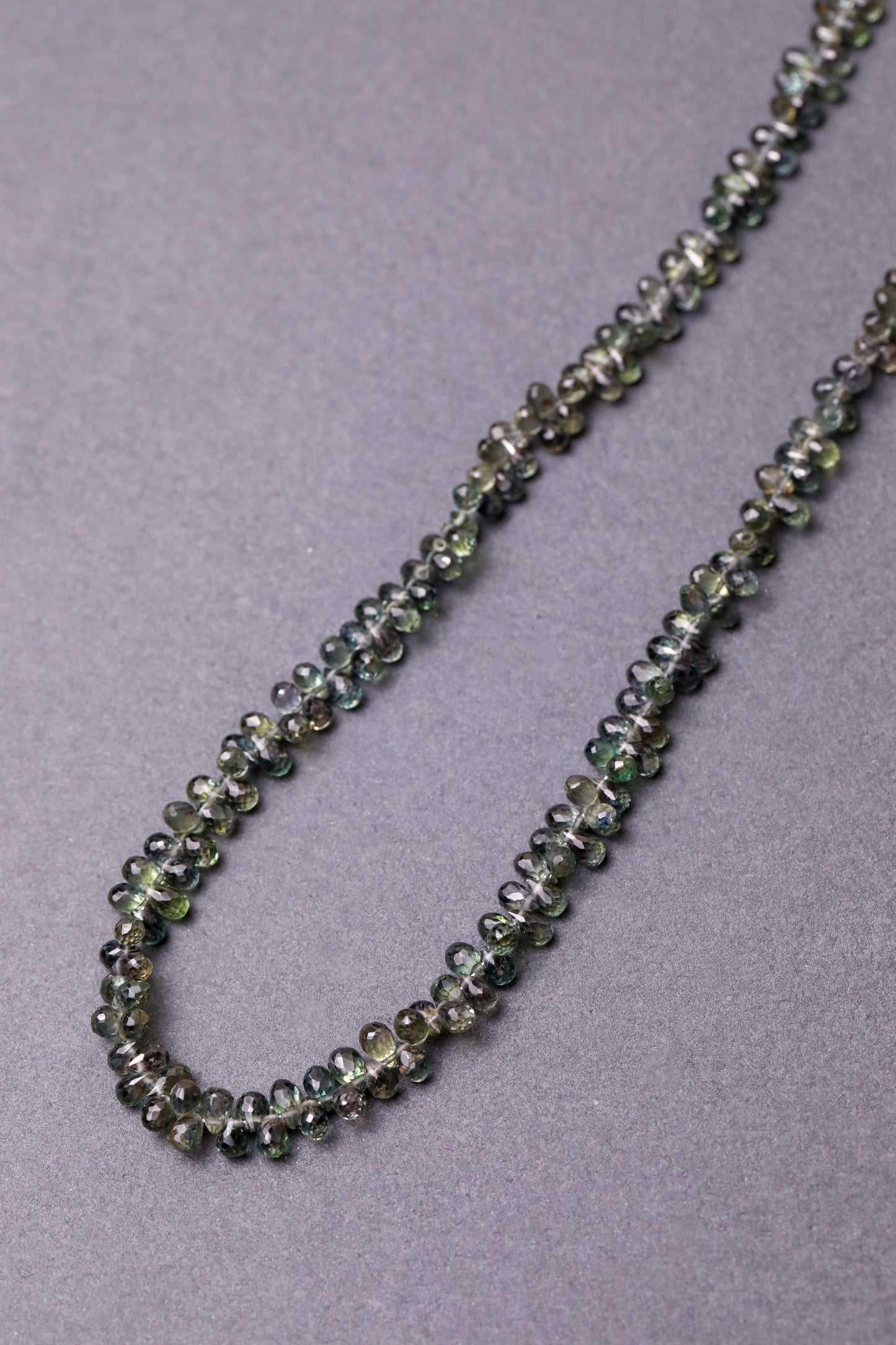 A necklace made of faceted teardrop-shaped Australian green sapphires, strung together  against a grey background.
