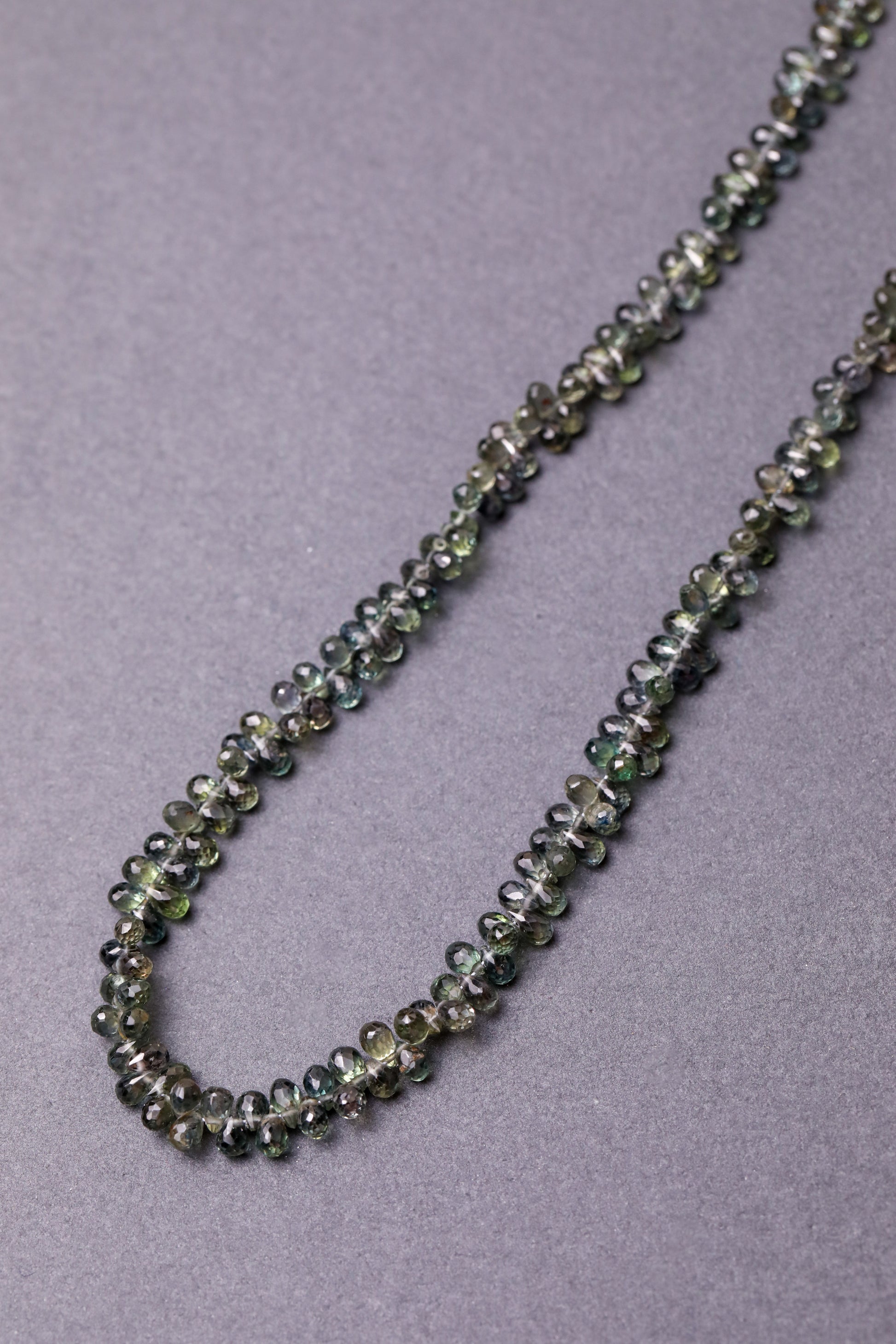 A necklace made of faceted teardrop-shaped Australian green sapphires, strung together  against a grey background.