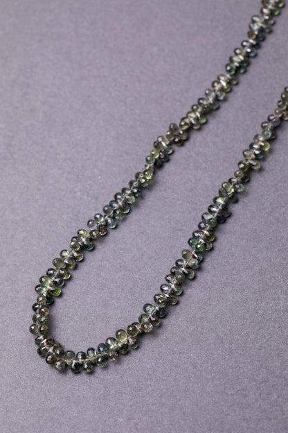 A necklace made of faceted teardrop-shaped Australian green sapphires, strung together  against a grey background.