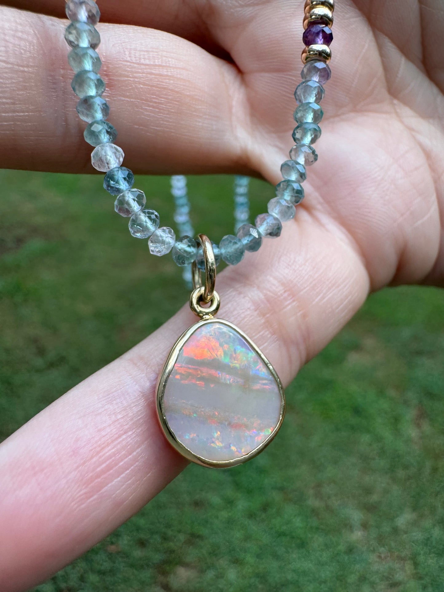 Close-up of a hand holding a beaded necklace with an opal pendant against a green background
