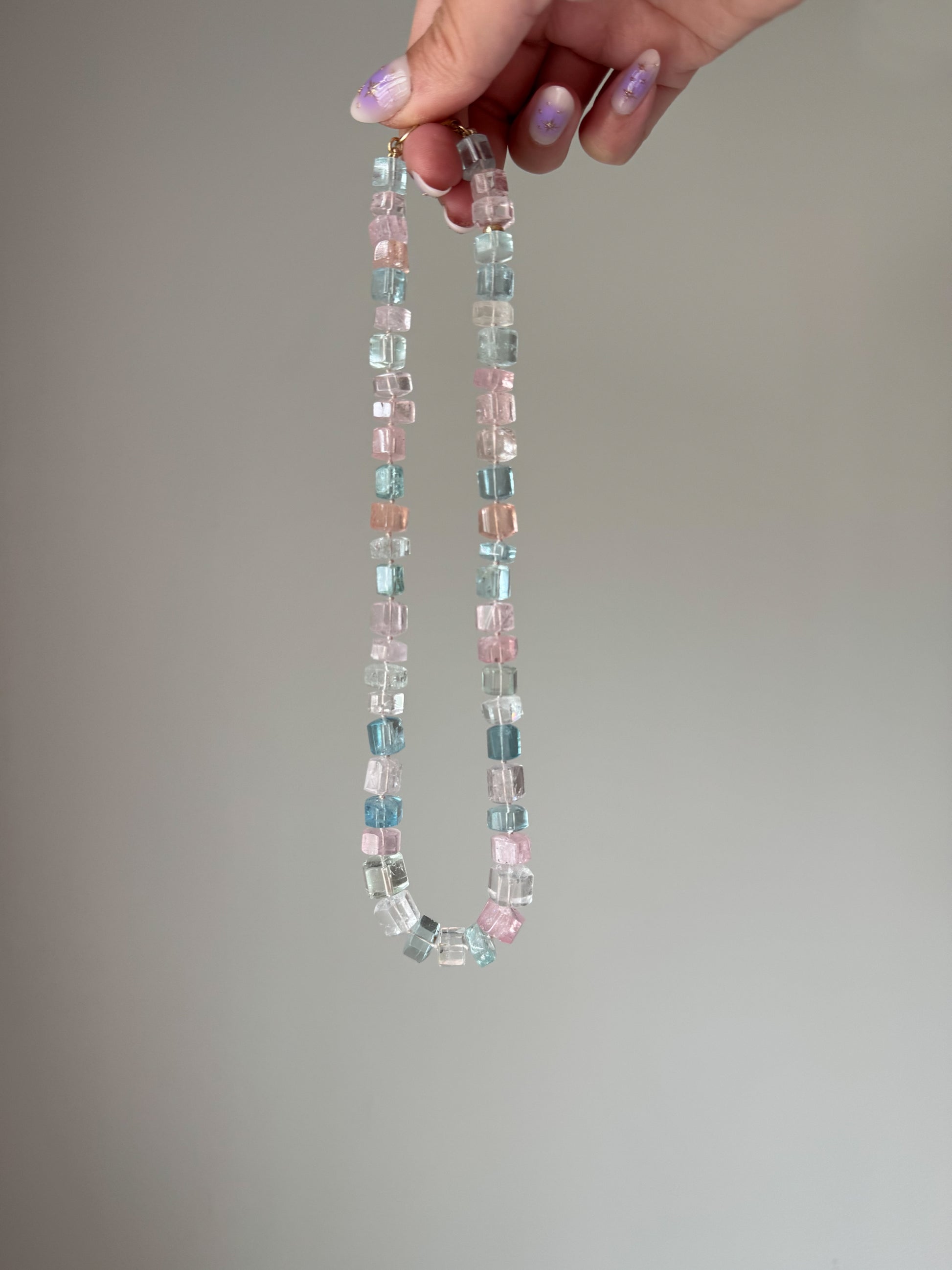 Necklace with multicolored gemstones held by a hand against a plain background. aquamarine pink blue green peach white 14k gold