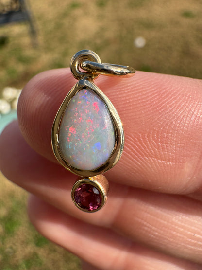 Gold pendant with opal and red gemstone held between fingers against a blurred natural background