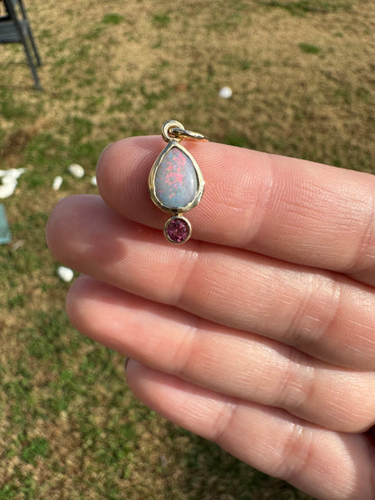 Gold charm with a teardrop-shaped gemstone and a small round gemstone on a finger against a blurred grassy background.