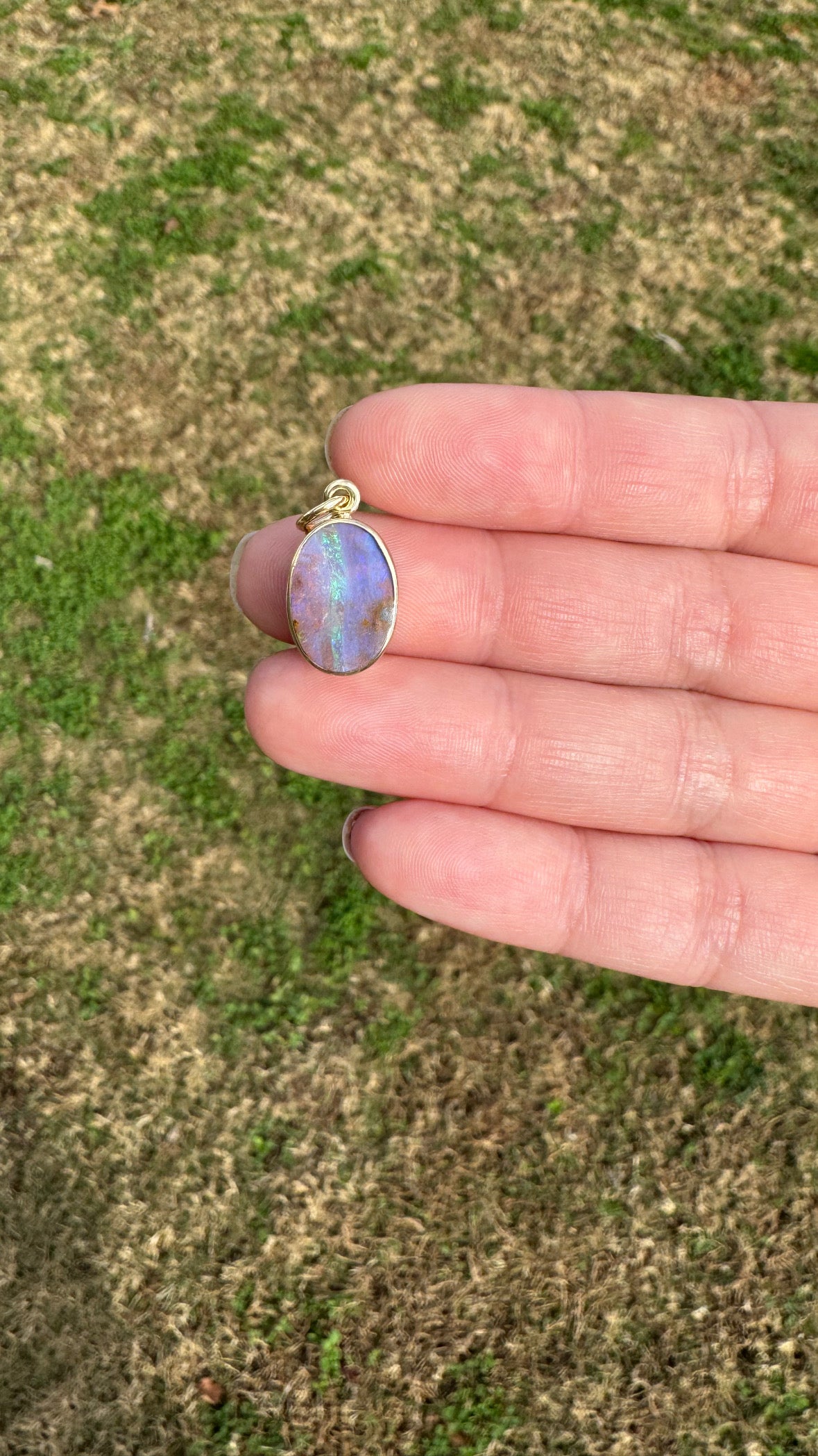 Hand holding a small oval pendant with a multicolored stone against a grassy background