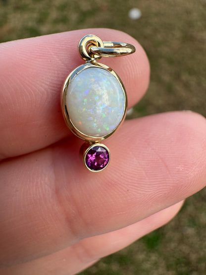 Gold pendant with opal and purple gemstone held between fingers against a blurred natural background