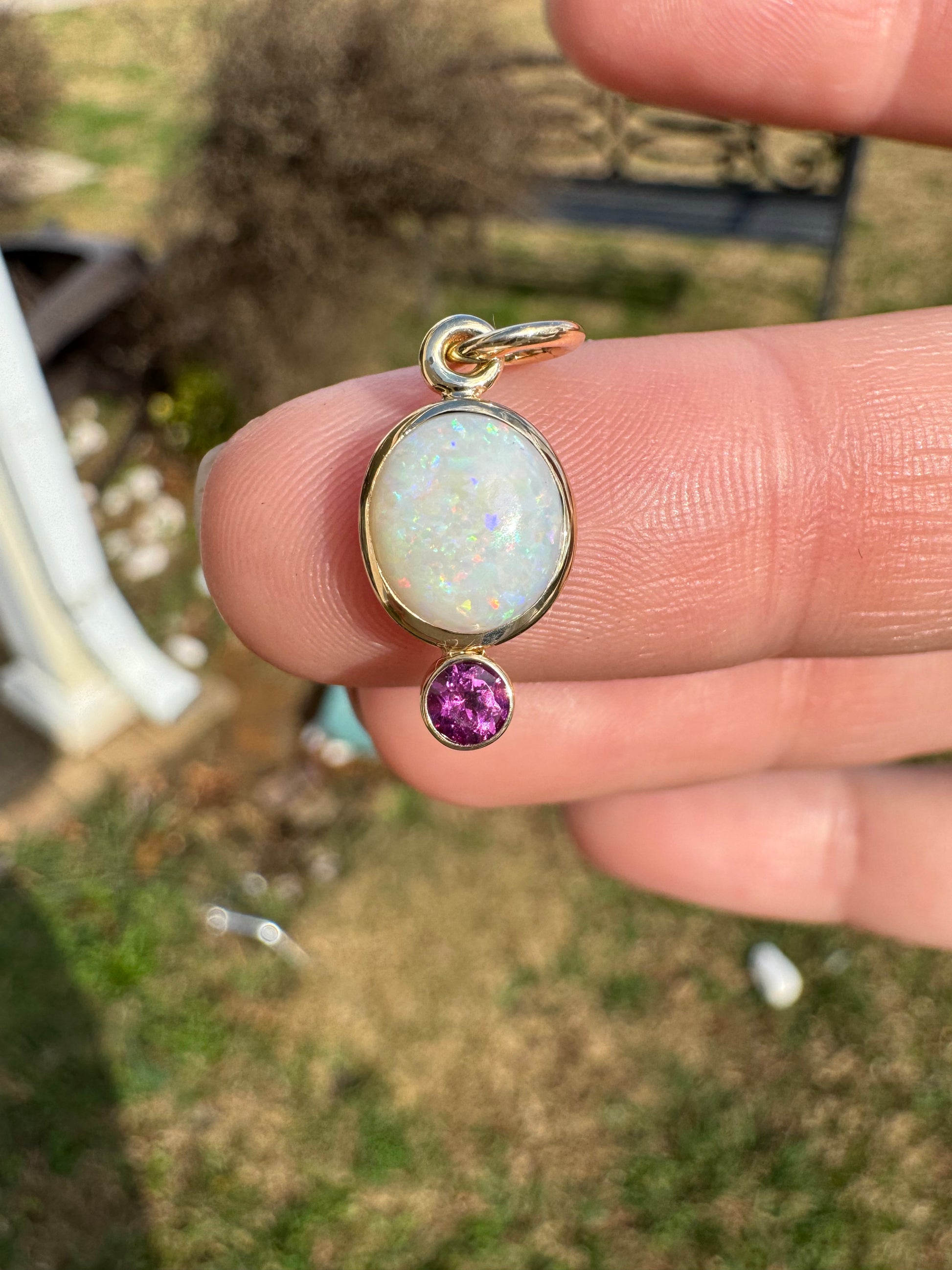 Gold pendant with opal and purple gemstone held between fingers against a blurred outdoor background