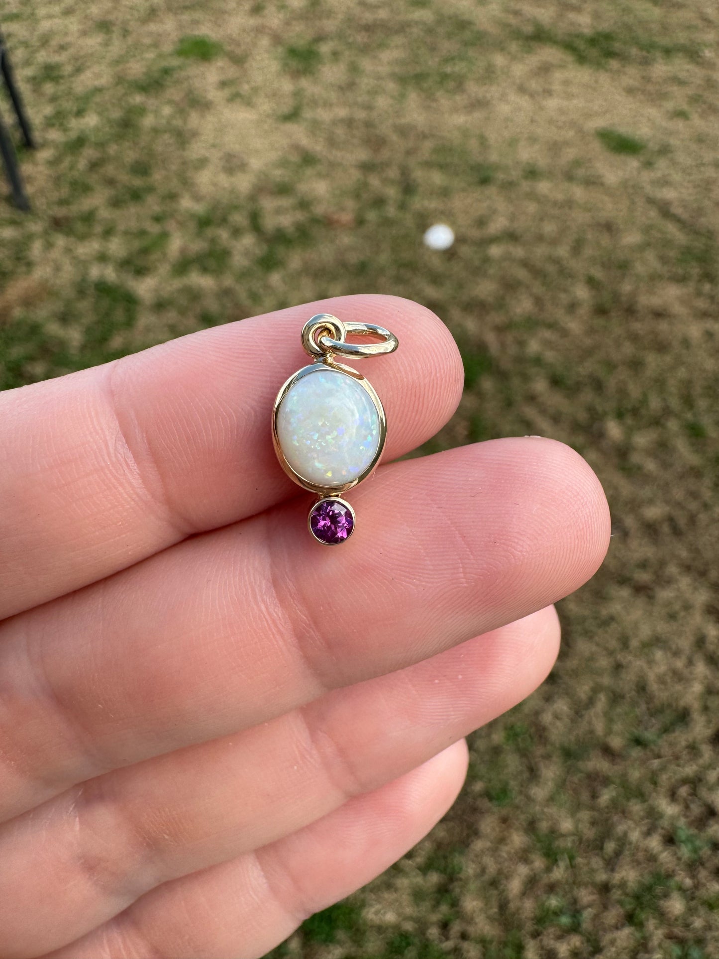 Gold pendant with opal and garnet stone held in a hand against a blurred natural background