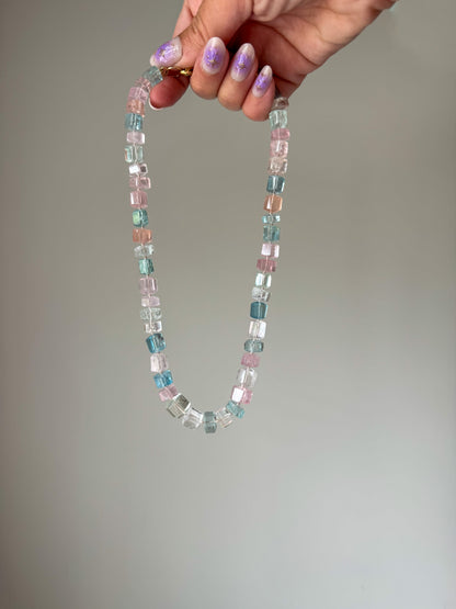 A knotted bead necklace with varying shades of aquamarine and light pink stones, held in a hand against a neutral background. aquamarine pink blue green peach white 14k gold