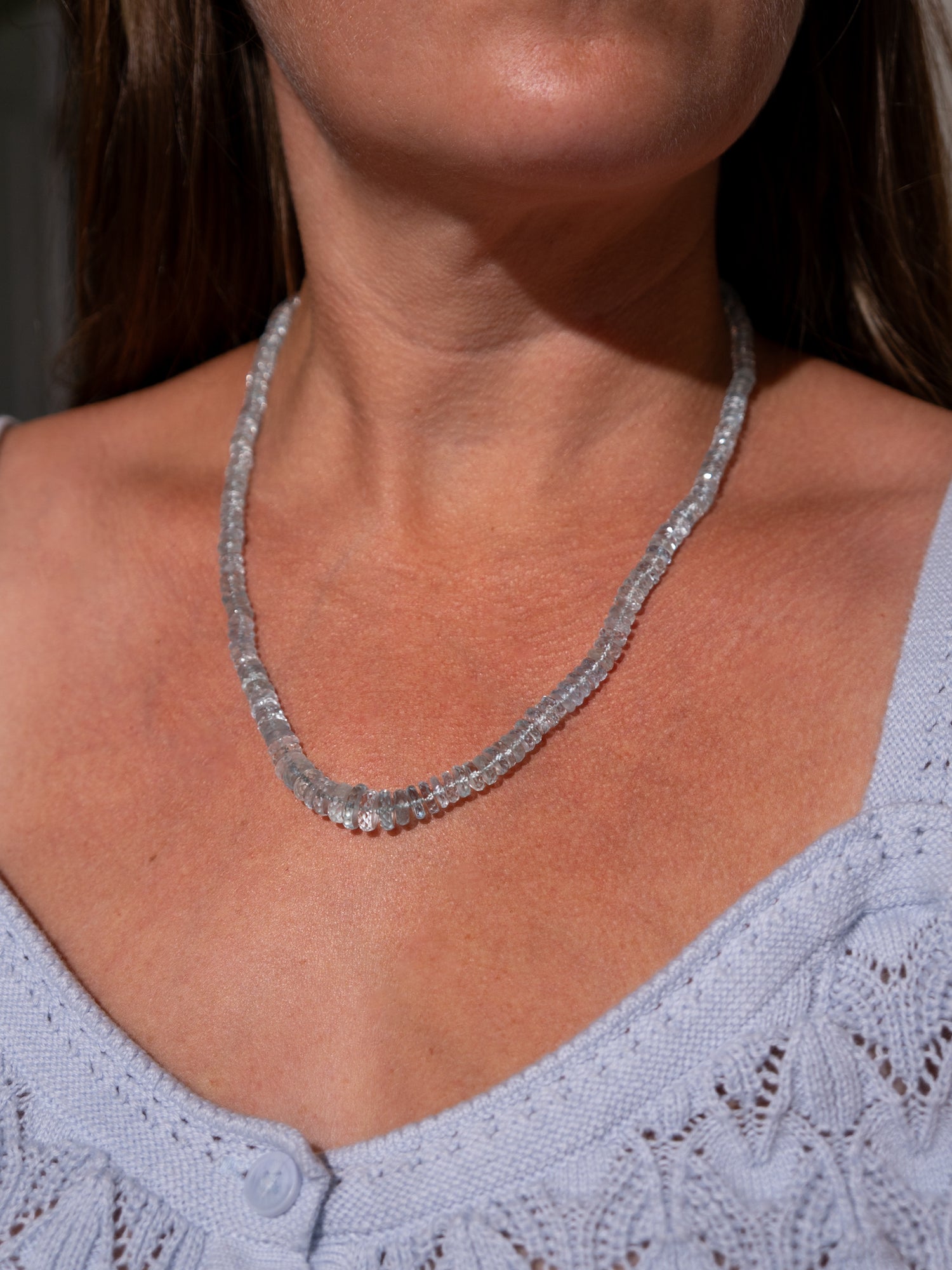 A close-up of a woman wearing an aquamarine faceted knotted heishi 14k gold necklace with clear gemstones, paired with a light blue knit top.