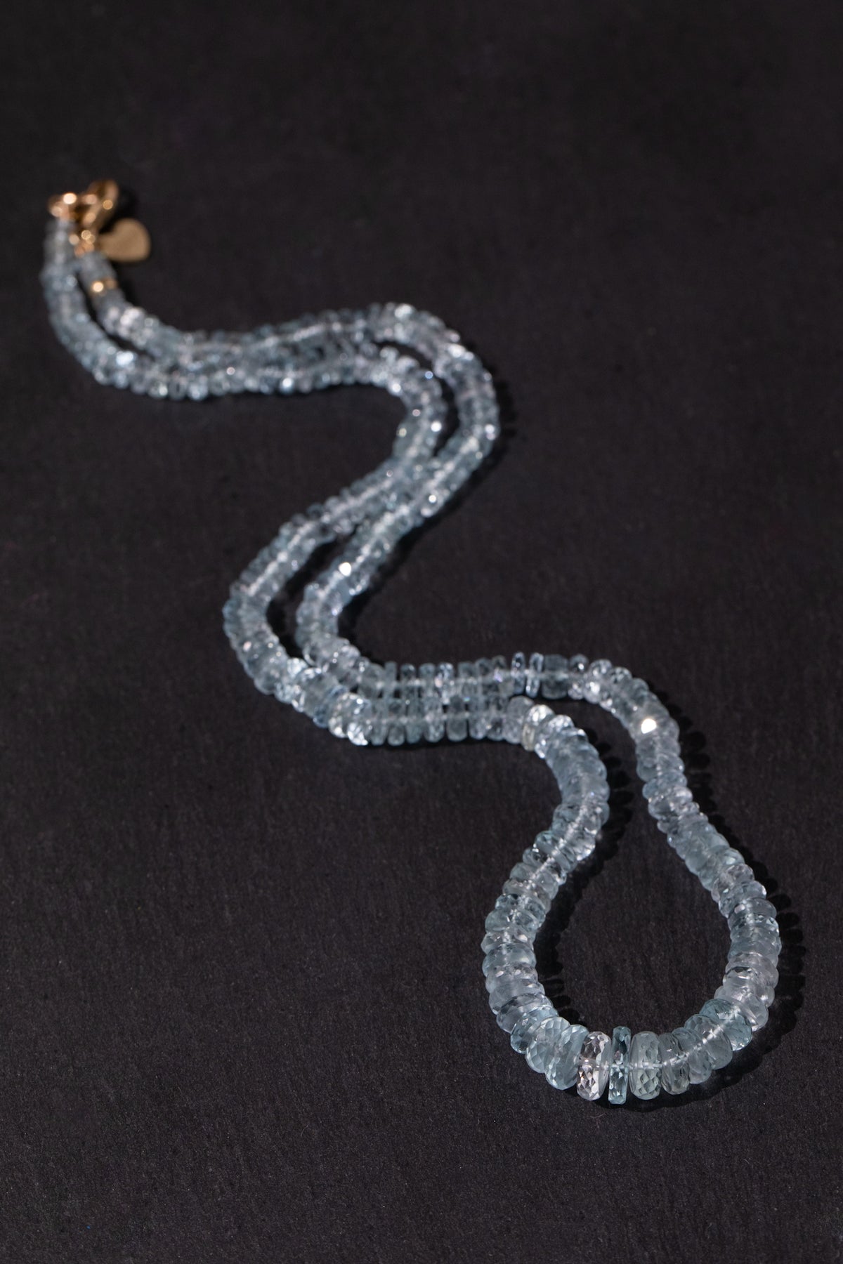 A double silk strand beaded necklace made of faceted light blue aquamarine beads, featuring a small gold heart charm and gold clasp, displayed on a dark textured background.