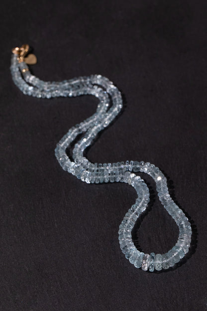 A double silk strand beaded necklace made of faceted light blue aquamarine beads, featuring a small gold heart charm and gold clasp, displayed on a dark textured background.