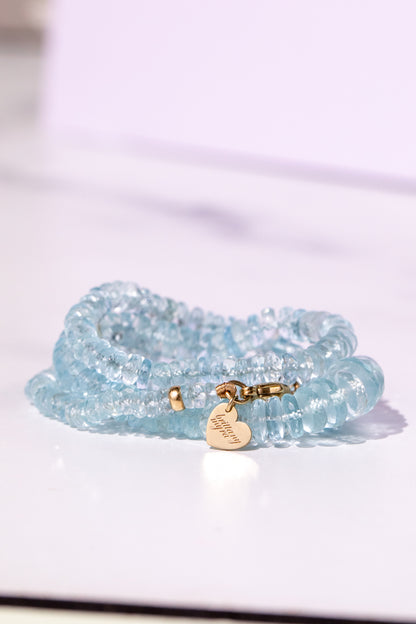 A light blue beaded long aquamarine necklace with a small gold heart charm engraved with 'brittany myra', resting on a white surface with a soft lavender background. silk knotted aquamarine candy necklace