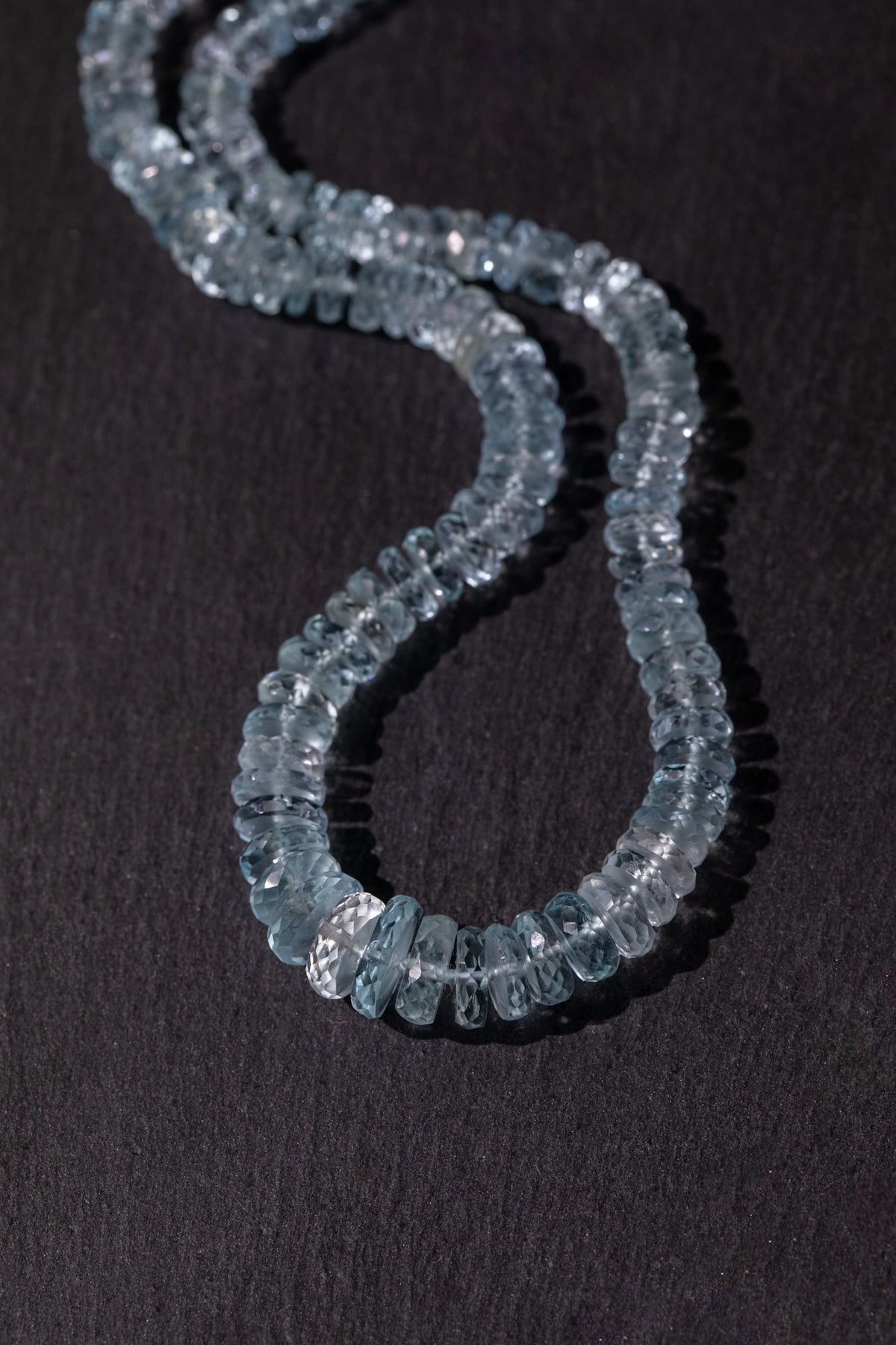 A beaded necklace made of faceted light blue gemstone aquamarine, arranged in a curved S-shape on a dark textured background.