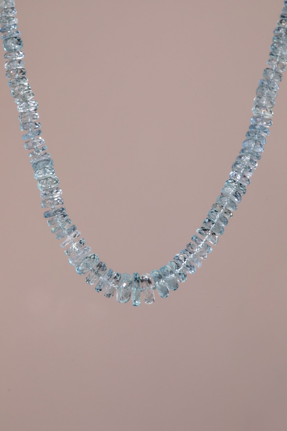 A delicate necklace made of scrumptious, light blue faceted  aquamarine gemstones, arranged in a single curved row, set against a solid pale pink background.