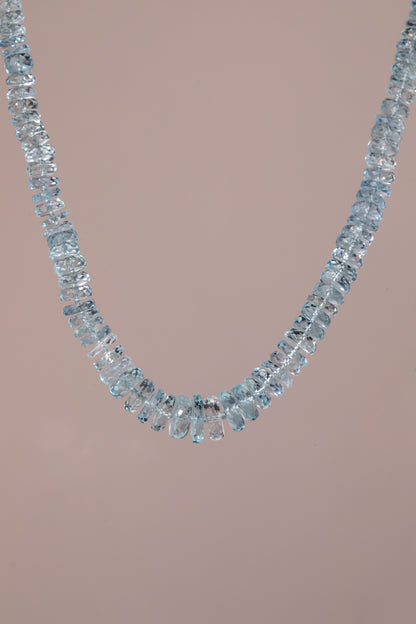 A delicate necklace made of scrumptious, light blue faceted  aquamarine gemstones, arranged in a single curved row, set against a solid pale pink background.