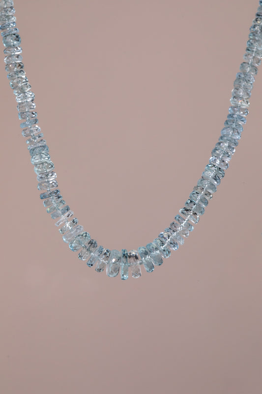 A delicate necklace made of scrumptious, light blue faceted  aquamarine gemstones, arranged in a single curved row, set against a solid pale pink background.