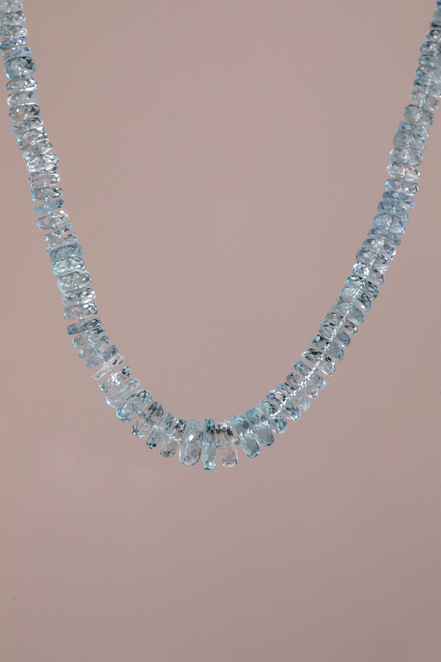 A delicate necklace made of scrumptious, light blue faceted  aquamarine gemstones, arranged in a single curved row, set against a solid pale pink background.