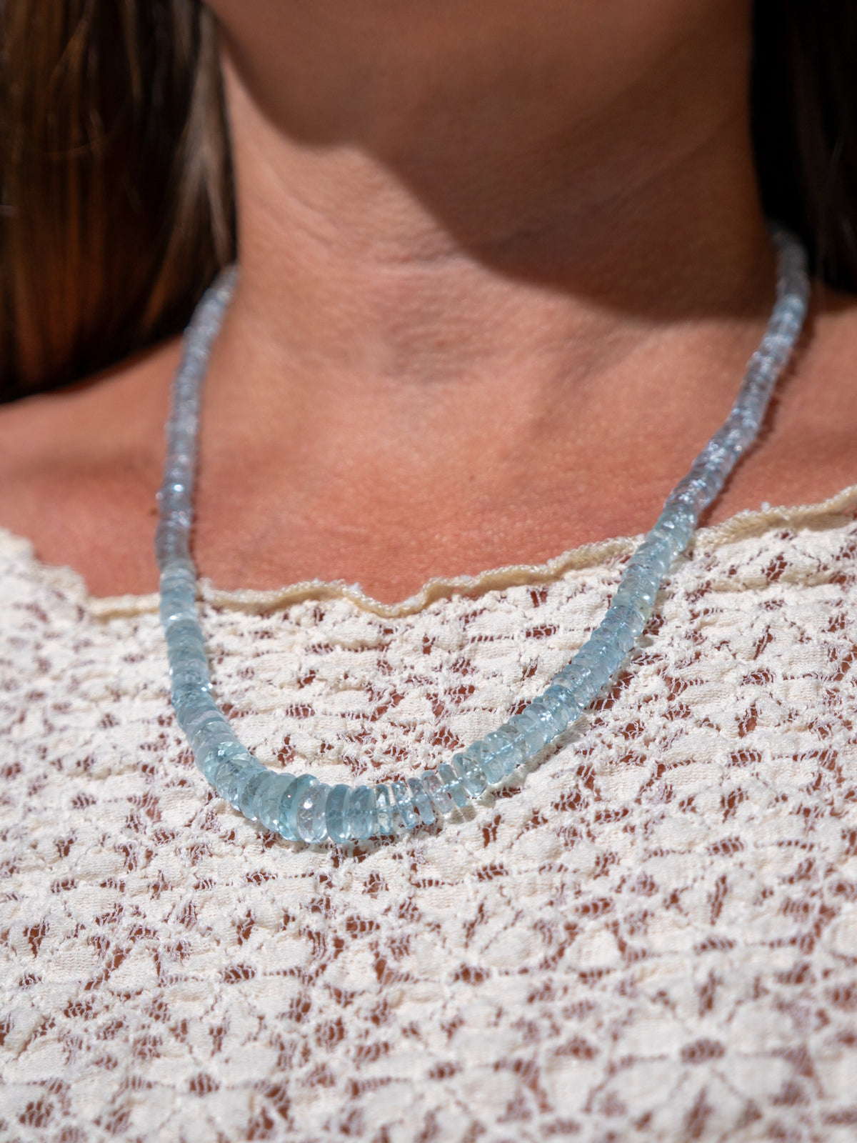 A close-up of a person wearing a light blue faceted aquamarine gemstone beaded necklace over a cream lace top.