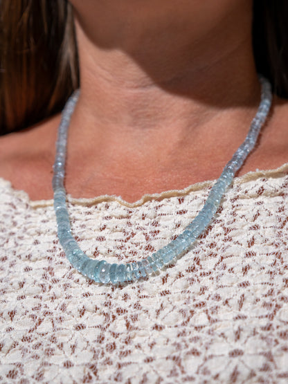 A close-up of a person wearing a light blue faceted aquamarine gemstone beaded necklace over a cream lace top.