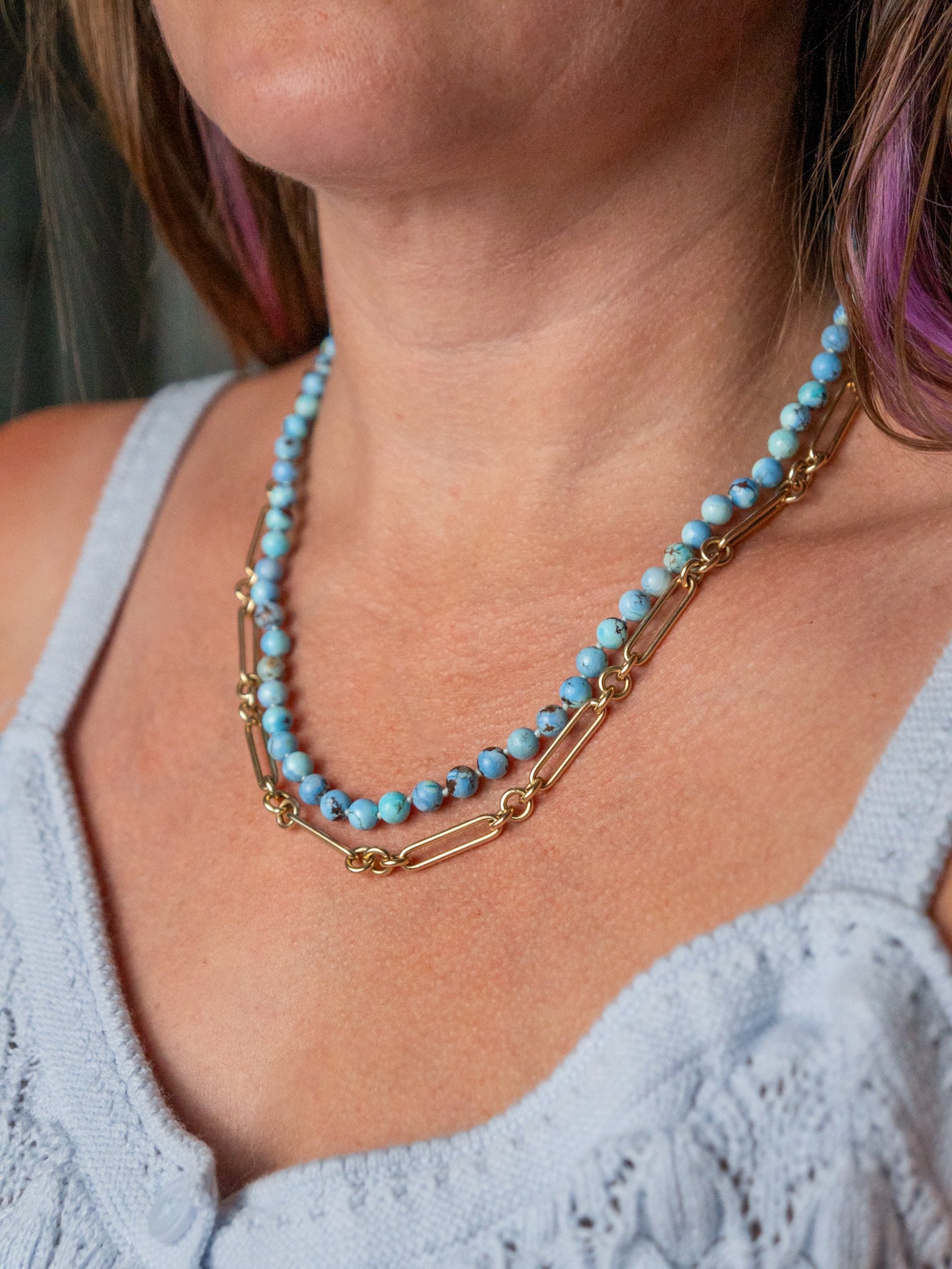 A close-up of a woman wearing a double-strand kazakhstan turquoise necklace with light blue beads and gold-toned chain links, paired with a light blue knit top.