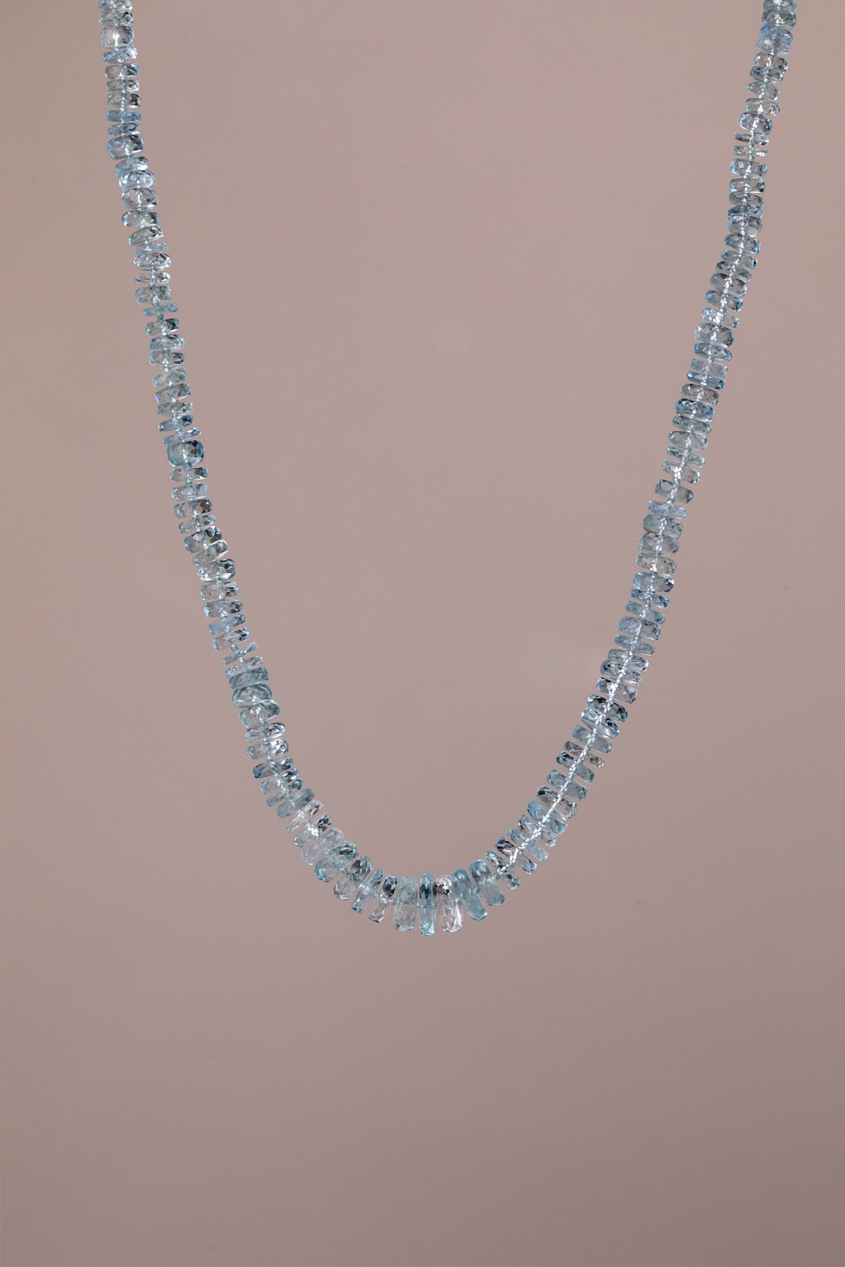A delicate necklace made of authentic aquamarine, faceted light blue gemstones, arranged in a curved V-shape against a solid, muted dusty pink background.