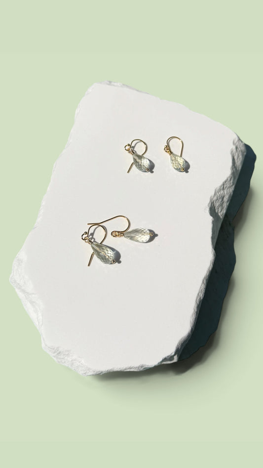 A pair of gold-toned drop earrings with clear teardrop-shaped stones, displayed on a white textured stone slab against a pale green background. prasiolite gold filled